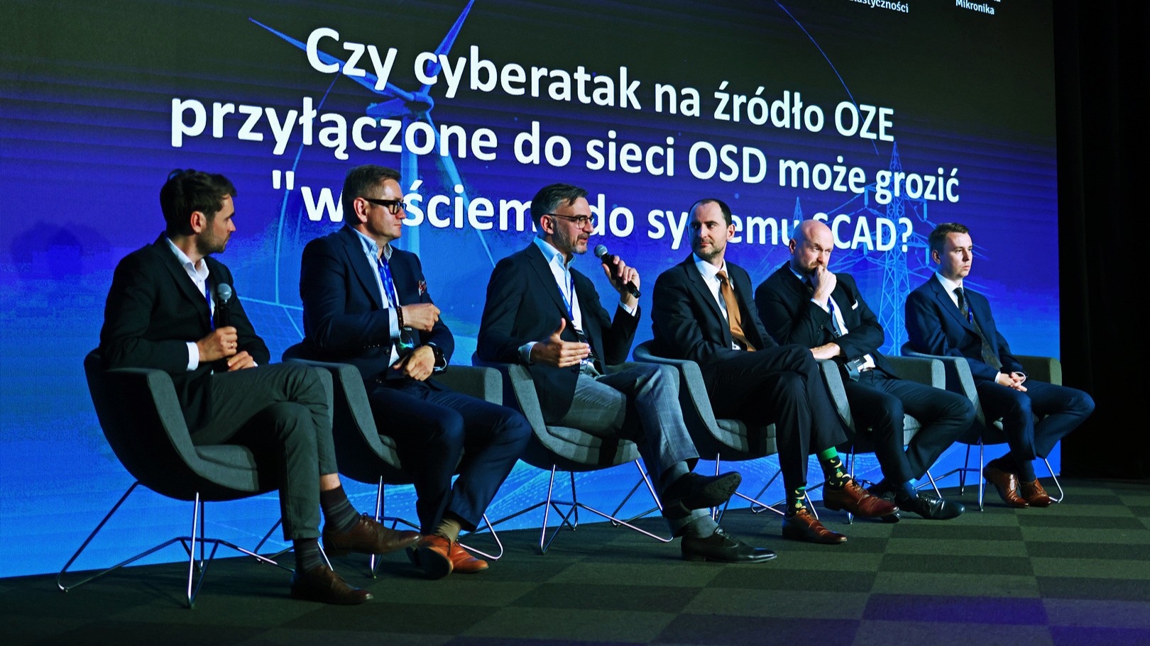 6 osób w garniturach siedzących na scenie na tle dużego ekranu LCD. 